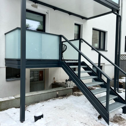 Schwarze Metalltreppe mit Glasgeländer zu einem modernen Balkon an einer weißen Hausfassade im Schnee