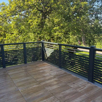 Holzterrasse mit dunklem Geländer und Blick auf üppiges grünes Baubestand im Hintergrund