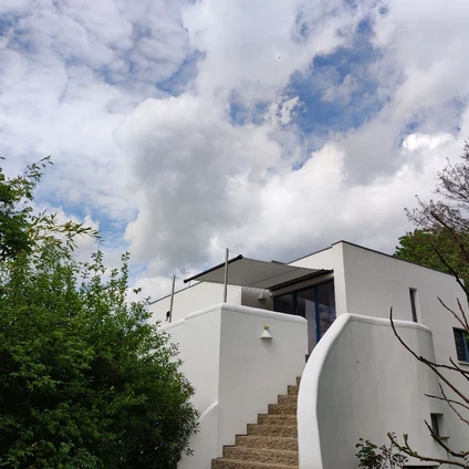 Modernes weißes Wohnhaus mit Treppe vor einem Wolkenhimmel und umgeben von üppigem Grün
